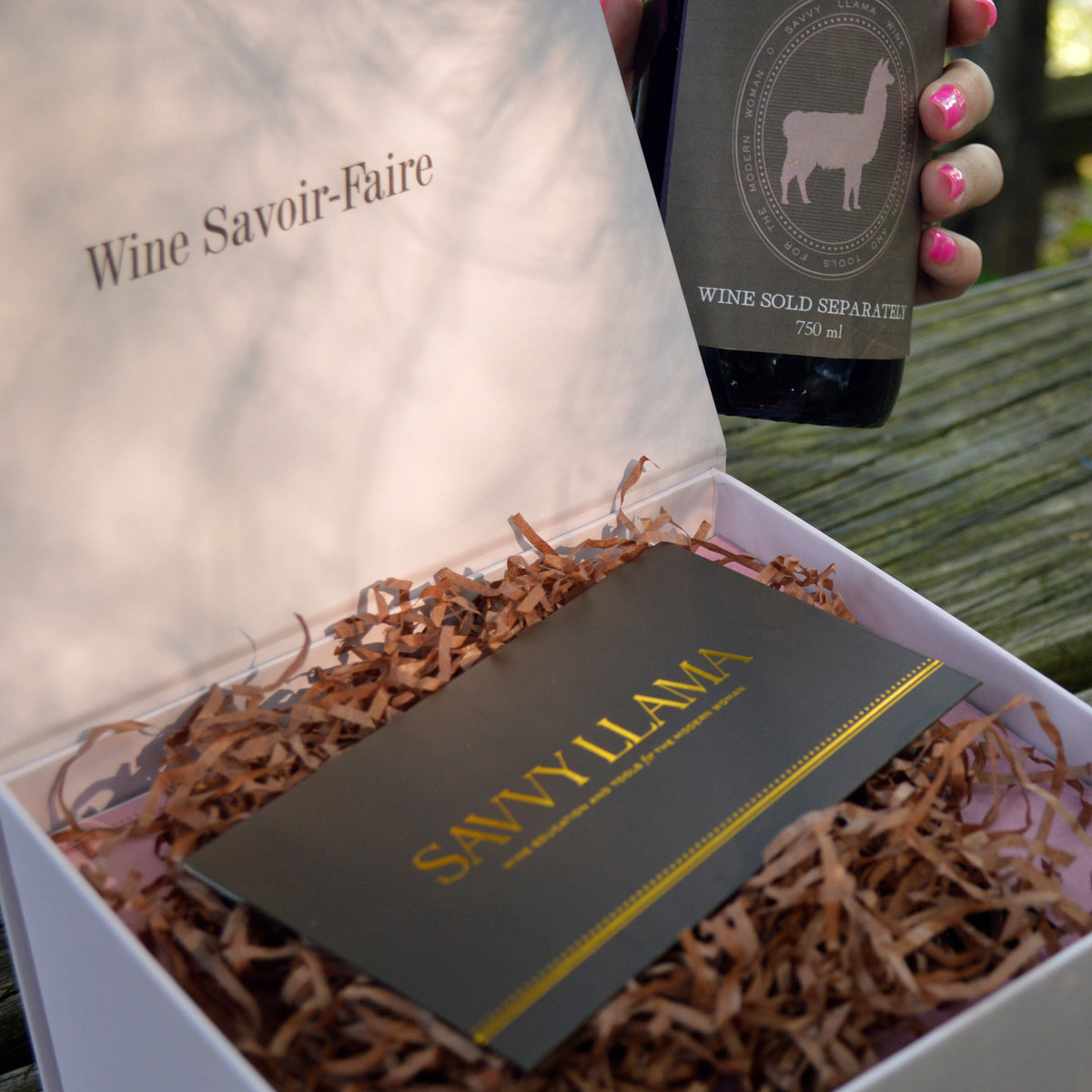 A Savvy Llama Savvy Decanting Kit elegant light pink gift box with gold foil lettering reads "Wine Savoir-Faire" on th inside lid, opened on a wooden table, containing a dark wine bottle held by a hand with pink nails, a branded card, and shredded brown paper filler. 