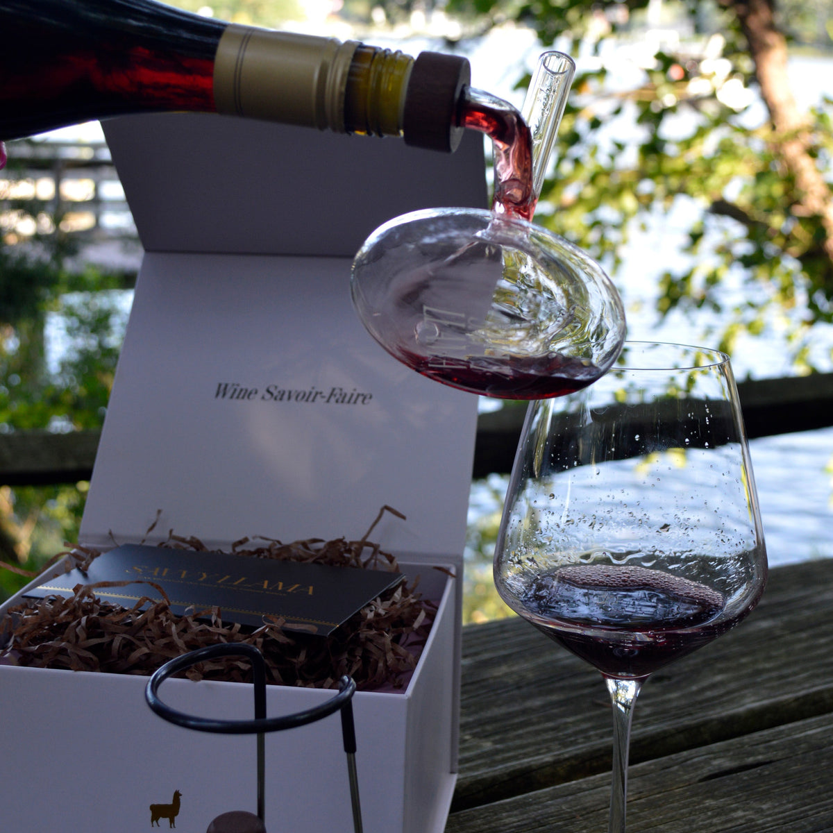 A Savvy Llama glass on-the-bottle mini wine decanter sits atop a bottle of red wine being poured into a glass as it aerates, with an open pink gift box with gold foil, labeled 'Wine Savoir-Faire' on a wooden table outdoors. A filled wine glass and its stainless steel decanter stand sits nearby, and trees and water are visible in the background.