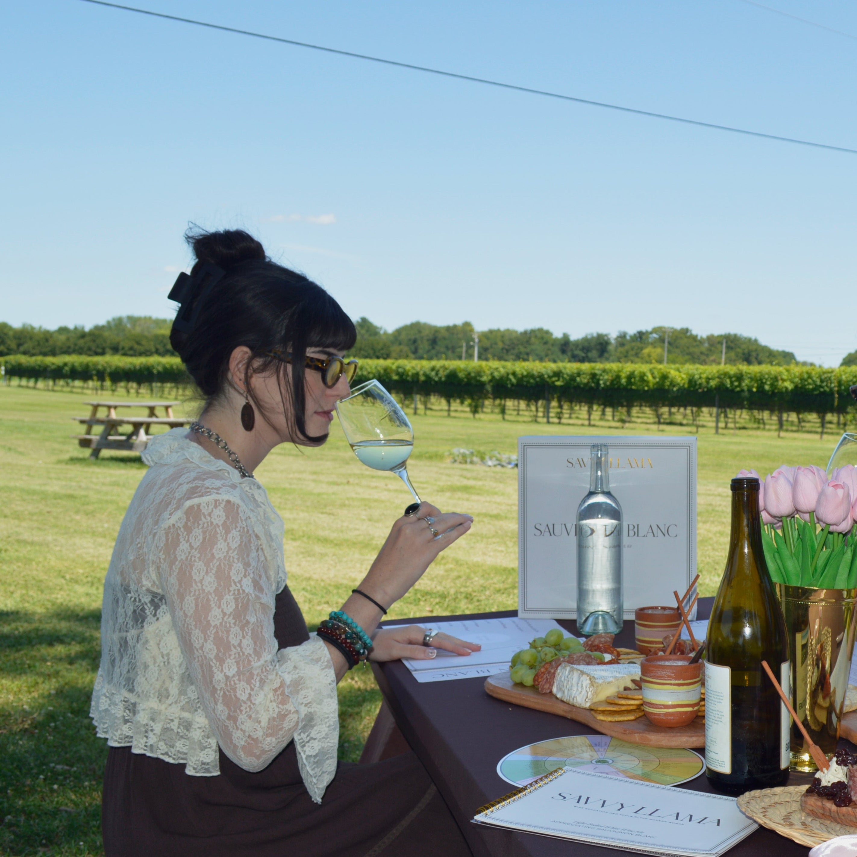 Brunette woman outdoors at a table in a vineyard setting, holding a wine glass, practicing a wine tasting ritual. The table has wine bottles, food, papers, and a vase of pink flowers.