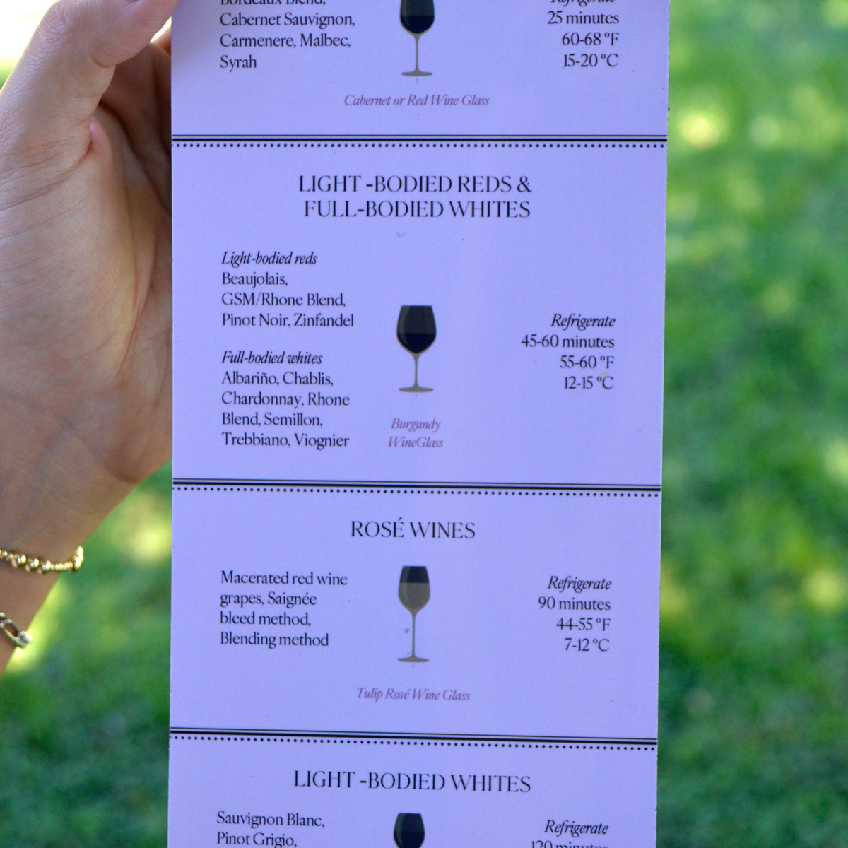 A hand holds a tall, narrow printed magnet with wine serving information, including text sections for full-bodied reds and whites, light-bodied reds and whites, rosé wines, with wine glass icons and temperature notes. The background is a blurred outdoor green scene.