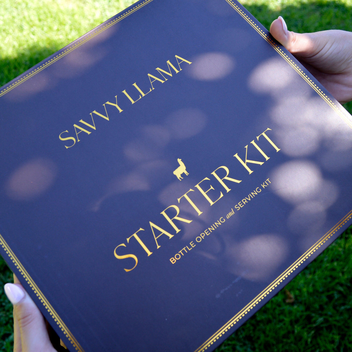 A person holds a dark Savvy Llama Starter Kit, a dark chocolate color box labeled 'SAWYILAMA + STARTER KIT BOTTLE OPENING AND SERVING KIT' with gold lettering, against a background of green grass with dappled sunlight.